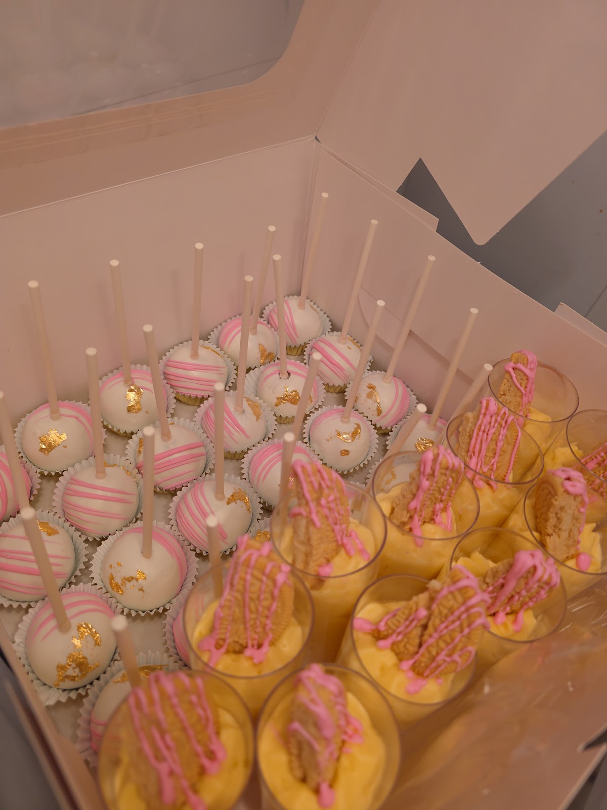 Boxed pink and white cake pops finished with gold leaf, beside banana pudding cups